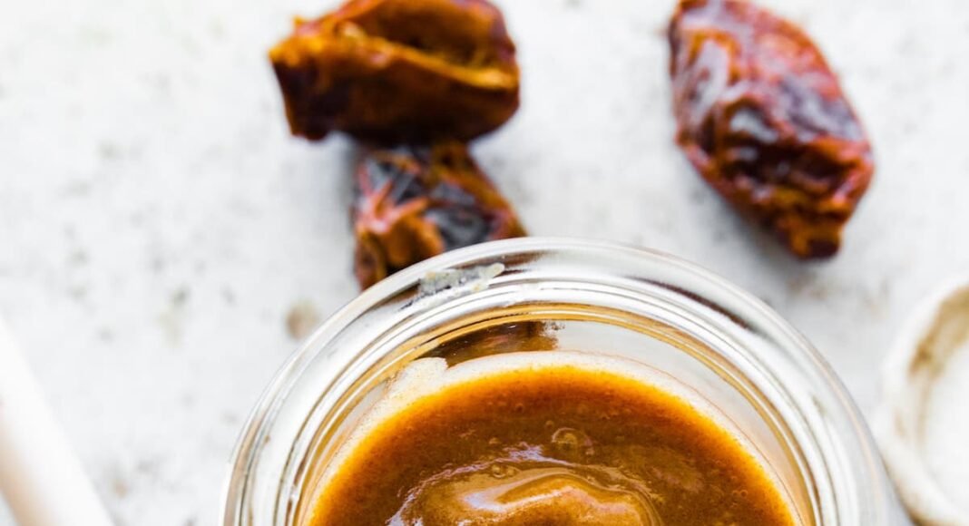 A glass jar of date caramel near a wooden spoon.