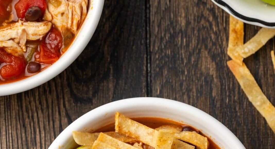 Overhead view of a bowl of chicken tortilla soup garnished with lime wedges and crispy tortilla strips.