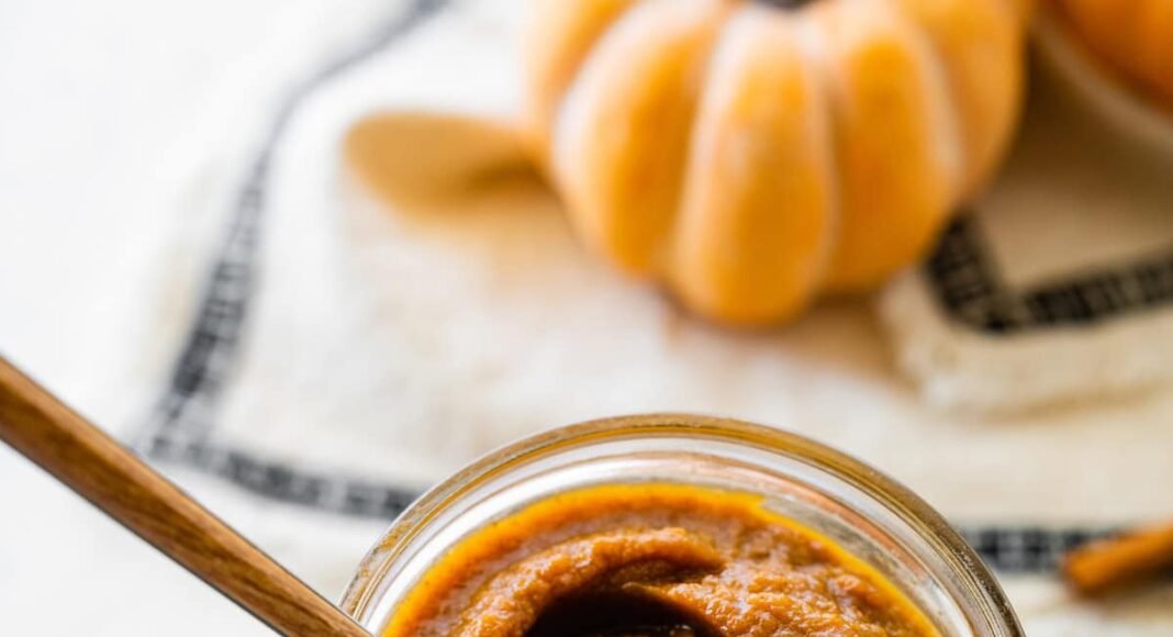 A glass jar full of pumpkin butter with a spoon inside.
