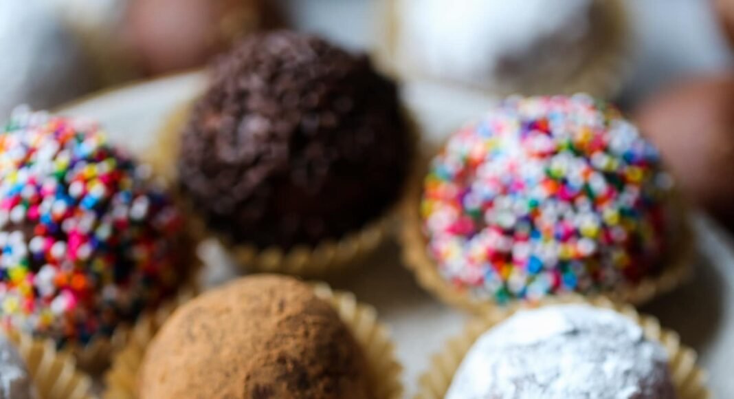 Decorated holiday rum balls in candy cups, arranged on a plate, one with a bite missing.