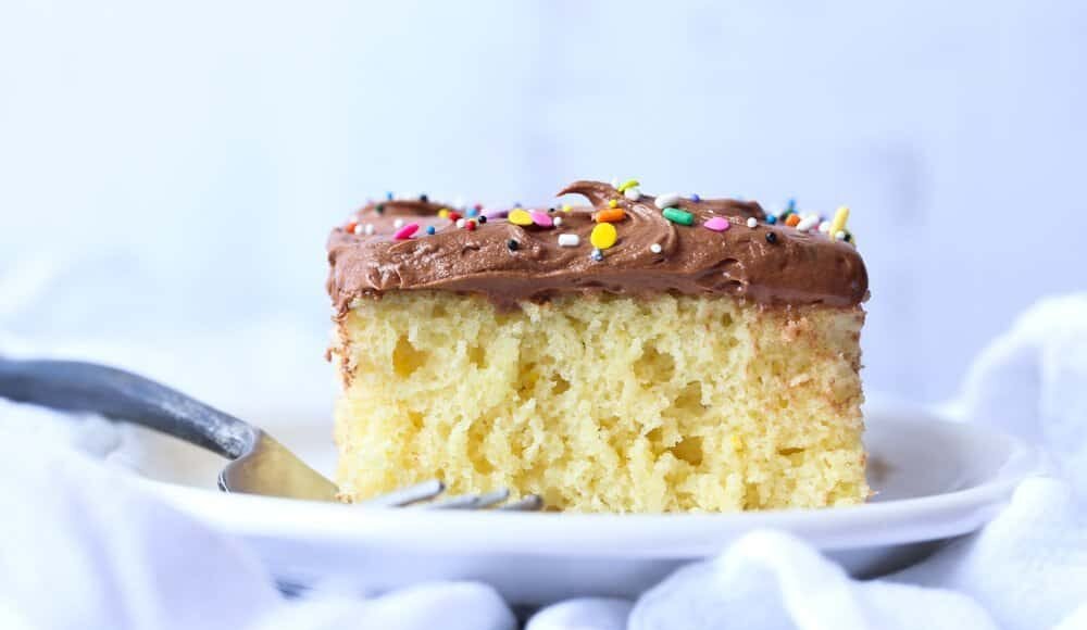Vanilla cake with chocolate frosting and rainbow sprinkles on a white plate with a fork.