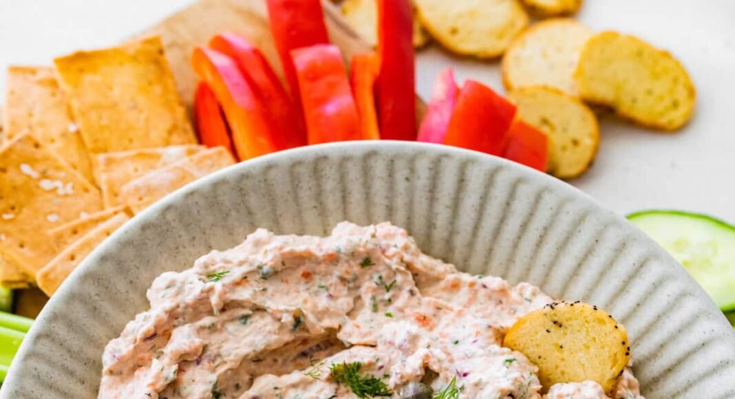 A bowl of smoked salmon dip, garnished with fresh dill and capers, served with crackers and fresh vegetables.