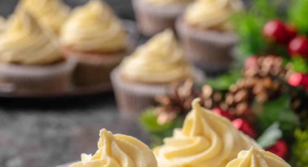 Three frosted gingerbread cupcakes on a white plate with Christmas decor and more cupcakes in the background.