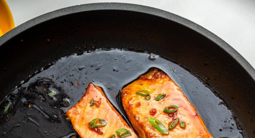 Four honey glazed salmon filets garnished with fresh chives in a skillet with a metal spoon that contains some of the honey based sauce in it.