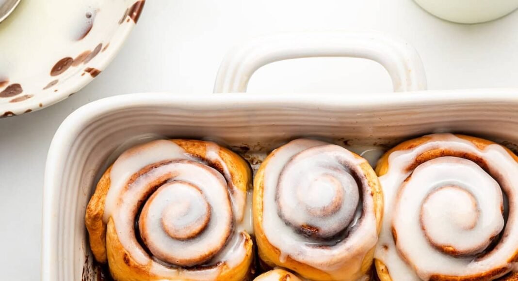 A white square baking dish filled with 9 baked cinnamon rolls that are topped with a glaze.