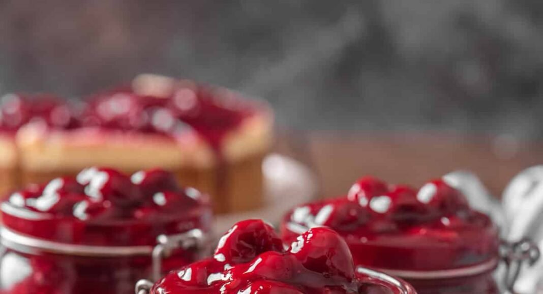 Three open jars of cherry pie filling on a round wooden tray, next to a spoonful of pie filling.