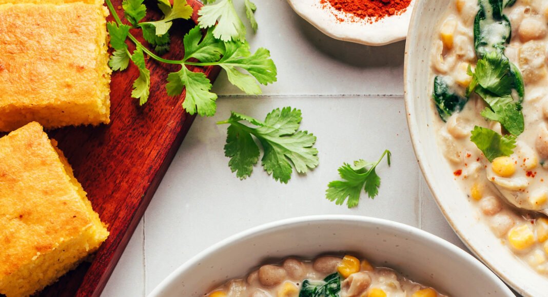 Bowls of creamy vegan white bean chili next to cayenne, cilantro, and cornbread