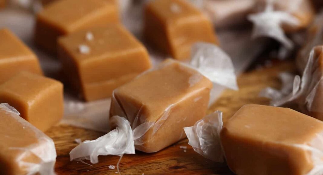 Homemade caramels wrapped in wax paper, arranged on a wood surface.