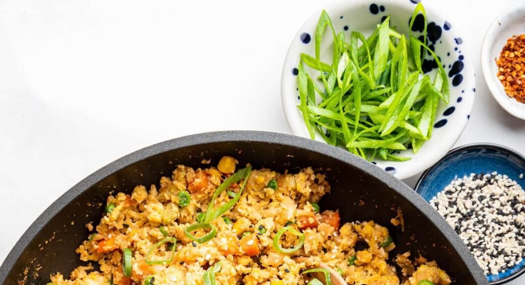 Cauliflower fried rice with eggs in a skillet with a wooden serving spoon. The dish is garnished with fresh green onion.