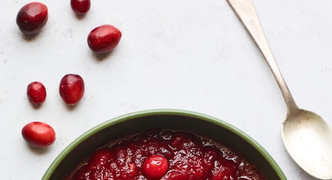 A serving dish containing cranberry sauce. A spoon rests at the top of the bowl and there are whole cranberries scattered around the bowl.