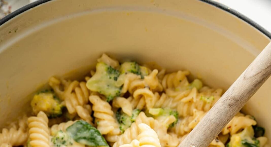 Rotini pasta, broccoli and spinach in a pot coated in a cheese sauce. A wooden spoon is scooping up some of the pasta.