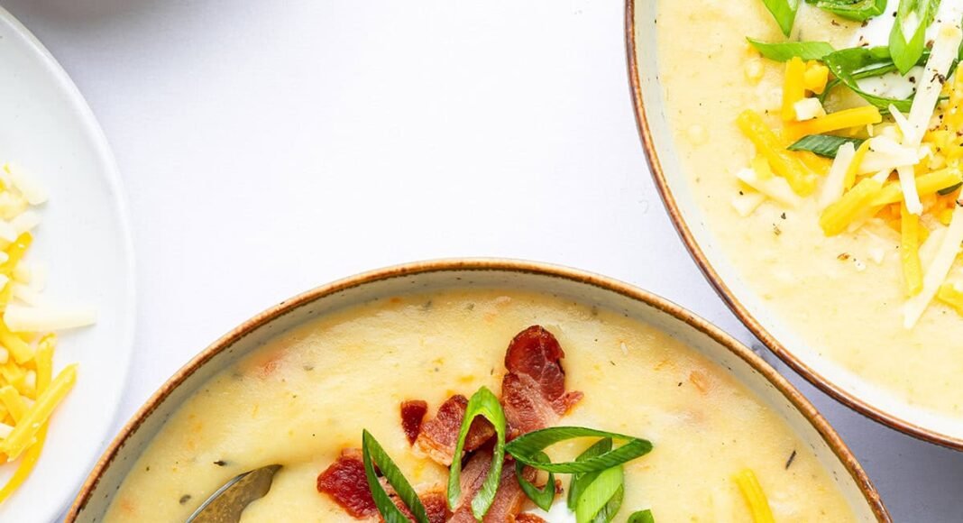 A bowl of potato soup with a silver spoon in it. The soup is topped with bacon bits, green onion, shredded cheese and sour cream.