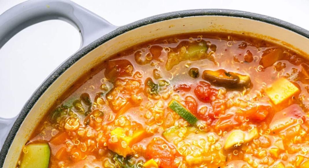 A large pot of vegetable packed quinoa soup with a wooden serving spoon in the soup.