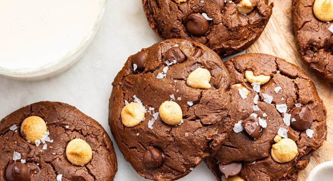 9 chocolate peanut butter cookies spread out on a counter. All of the cookies are topped with flaky sea salt.