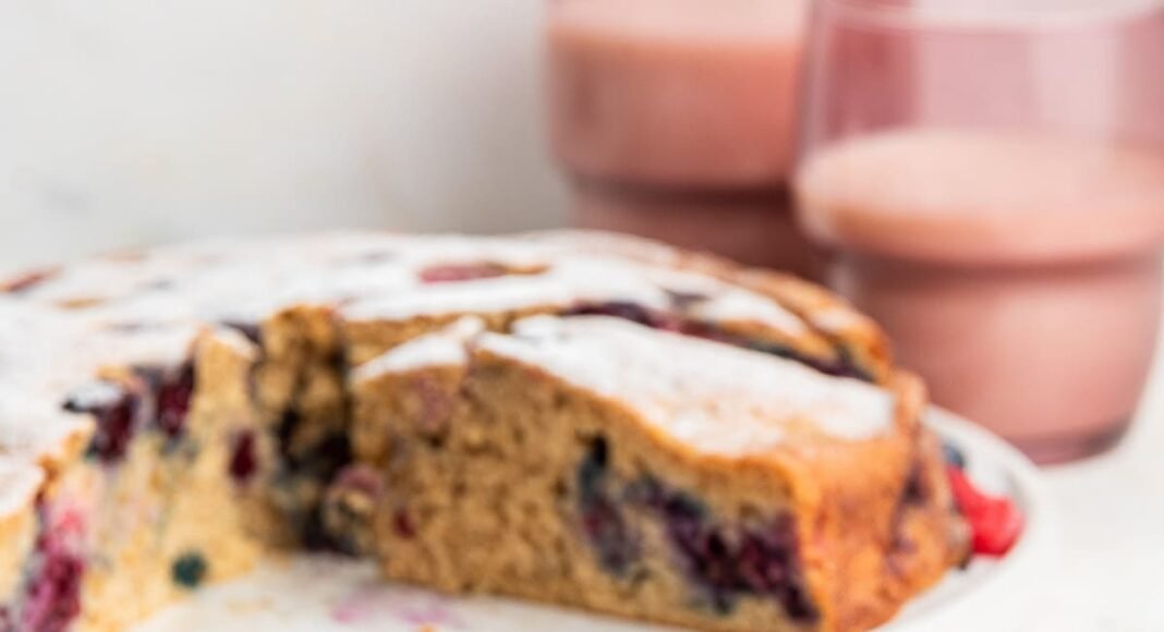 A slice of mixed berry cake on a light pink plate. The entire berry cake is in the background on a white plate.