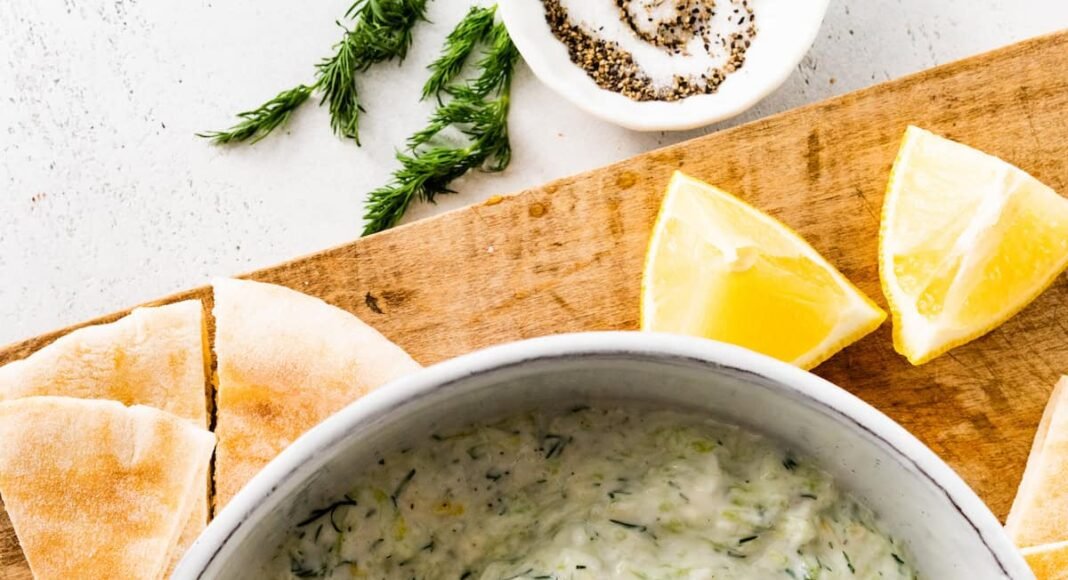 A white bowl of tzatziki topped with fresh dill. There is a wooden spoon in the bowl and the bowl is surrounded by pita bread and lemon slices.