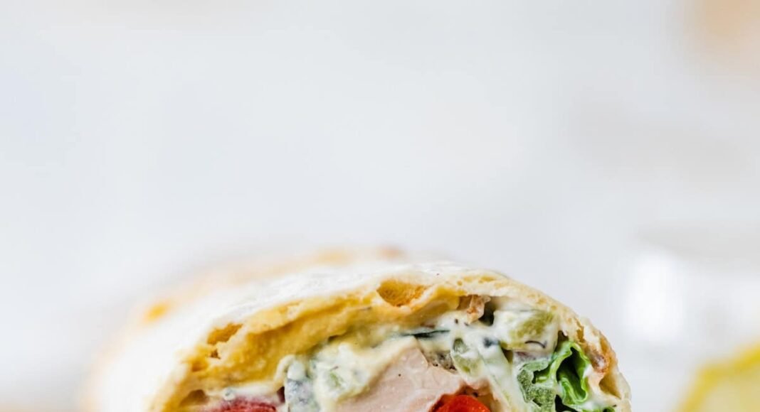 Two halves of a Greek chicken wrap stacked on top of each other on a wooden cutting board. You can see the tomatoes, chicken, lettuce and sauce inside the wrap.