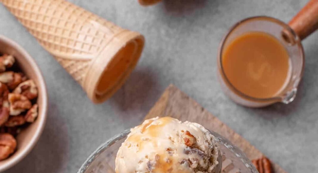 Overhead view of scoops of butter pecan ice cream in a glass dessert bowl, surrounded by ingredients.