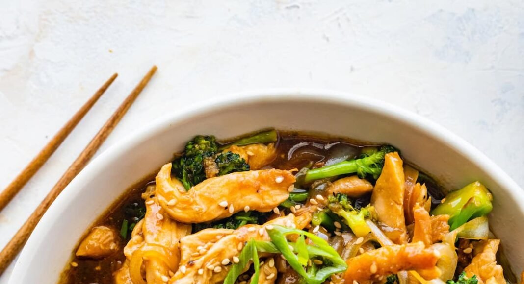 Chicken and broccoli stir fry in a white bowl, garnished with sesame seeds and green onions, with wooden chopsticks beside it.