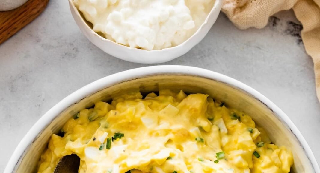 Cottage cheese egg salad served in a white ceramic bowl served with a silver spoon. Fresh chives are topping the egg salad.