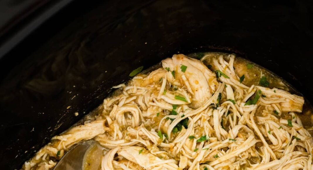 Shredded salsa verde chicken in a slow cooker with a silver serving spoon. Fresh cilantro is on top of the chicken.