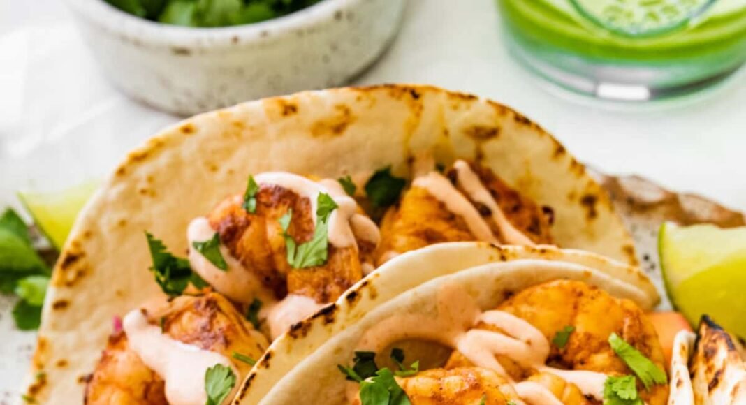 Three shrimp tacos in flour tortillas folded up on a serving platter. The tacos are topped with a mango cabbage slaw and a drizzle of spicy crema.