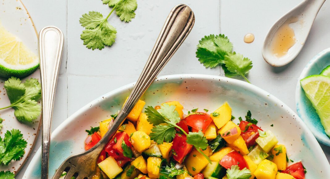 Fork and spoon resting in a bowl of Mango Cucumber Salad with lime wedges on top
