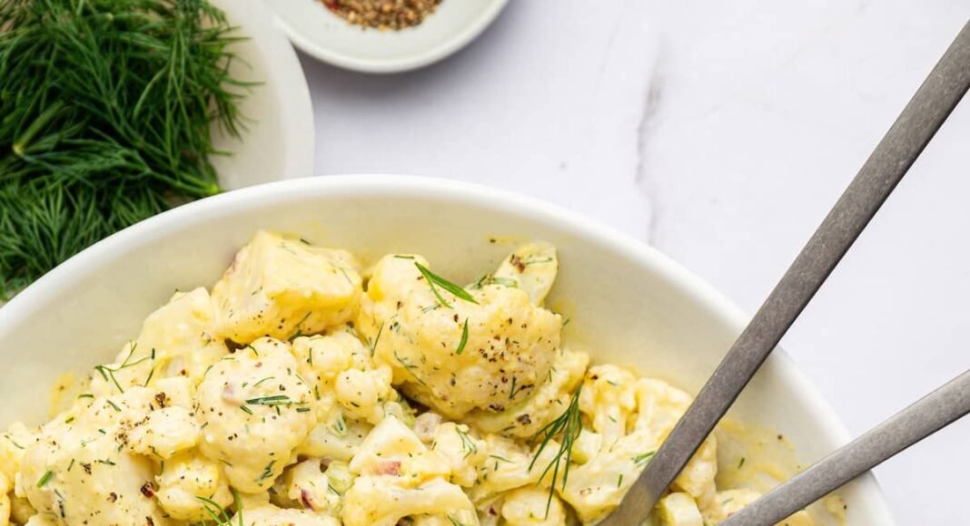 Cauliflower potato salad in a large serving bowl with silver serving utensils. The salad is topped with fresh dill.