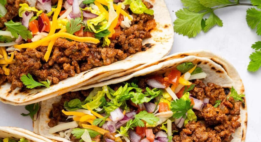 Five ground beef tacos in flour tortillas topped with lettuce, onion, tomato, cilantro and cheese.