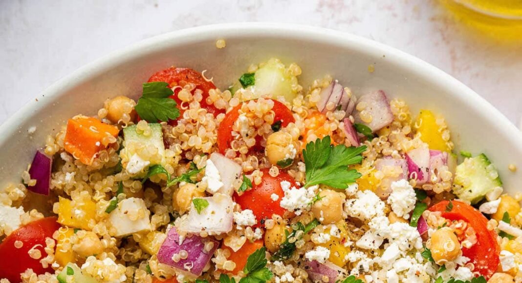 A mixed quinoa chickpea salad with tomatoes, cucumbers, feta, and parsley, in a white bowl.