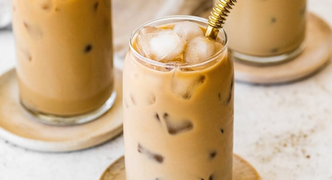 A glass of iced coffee with milk fully mixed in, served on a small wooden plate with a metal straw.