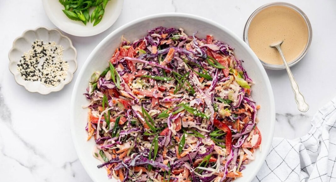 Assembled Asian slaw in a white bowl, garnished with sesame seeds and scallions. Dressing and toppings sit around the bowl on a marble counter with a striped towel.
