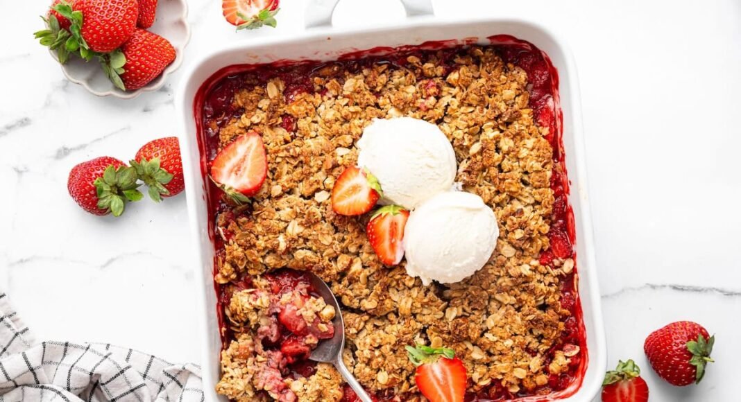 Baked strawberry crisp topped with two scoops of vanilla ice cream and fresh strawberry halves. There is a spoon scooping out some of the crisp.