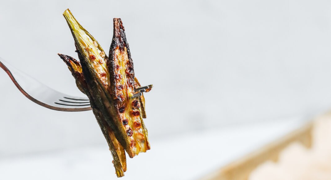 Fork holding up a few slices of crispy oven roasted okra over a plate of more baked okra