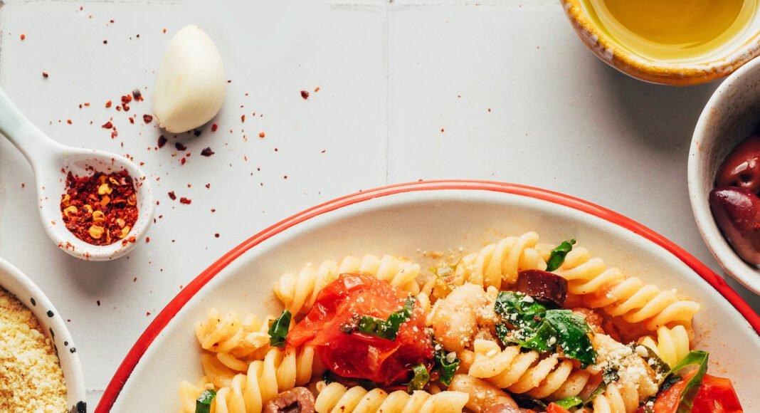 Large serving bowl of garlicky pasta with burst cherry tomatoes, kale, olives, and vegan parmesan cheese