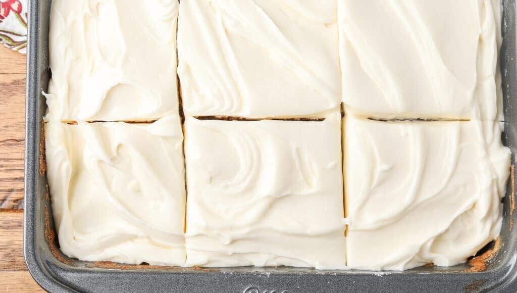 A horizontally aligned, top down close up photo of 6 slices of cake topped with fluffy cream cheese frosting in a metal pan.