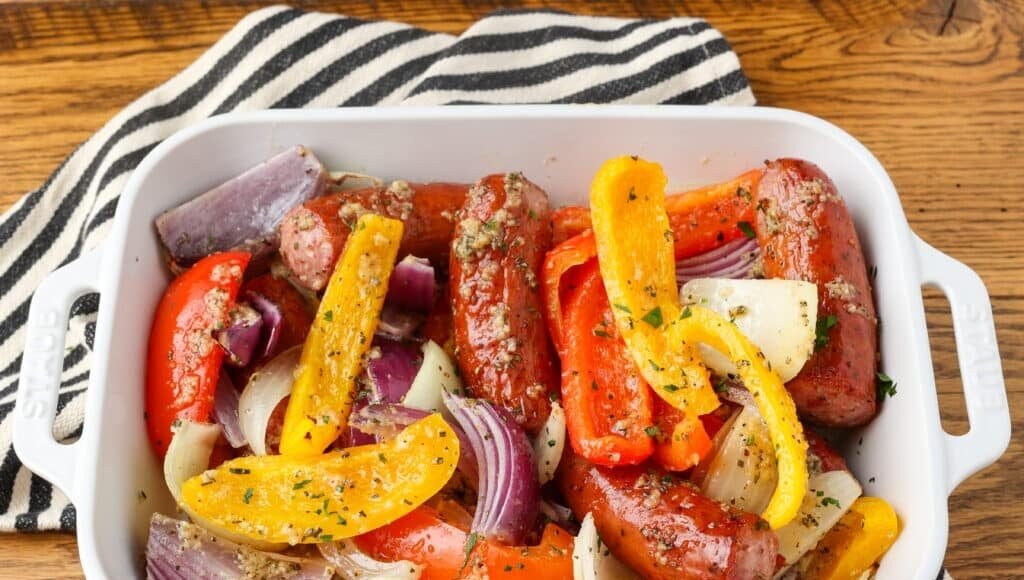 A close up shot of a baking dish filled with sausage and peppers with onions, over a black and white striped tea towel on a wooden tabletop.