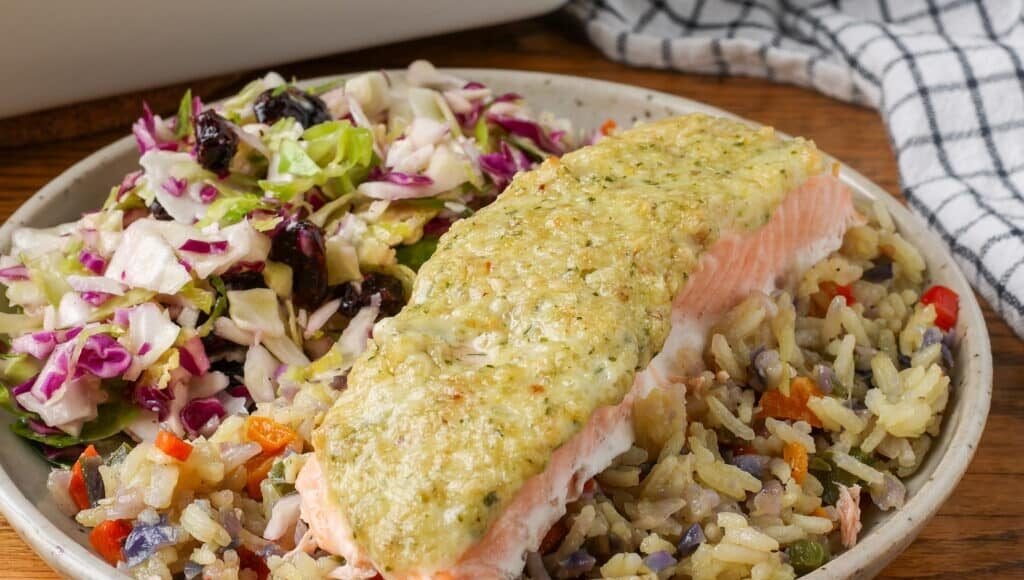 A horizontally aligned close up photo of the pesto salmon atop a bed of rice pilaf next to a serving of salad on a plate over a wooden table.